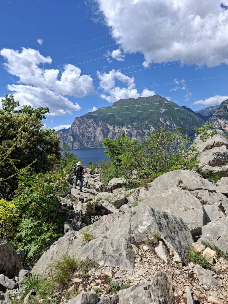 Wanderer auf einem felsigen Abschnitt des Busatte-Tempesta Wanderwegs oberhalb vom Gardasee mit Blick auf Berge, See und blauen Himmel.