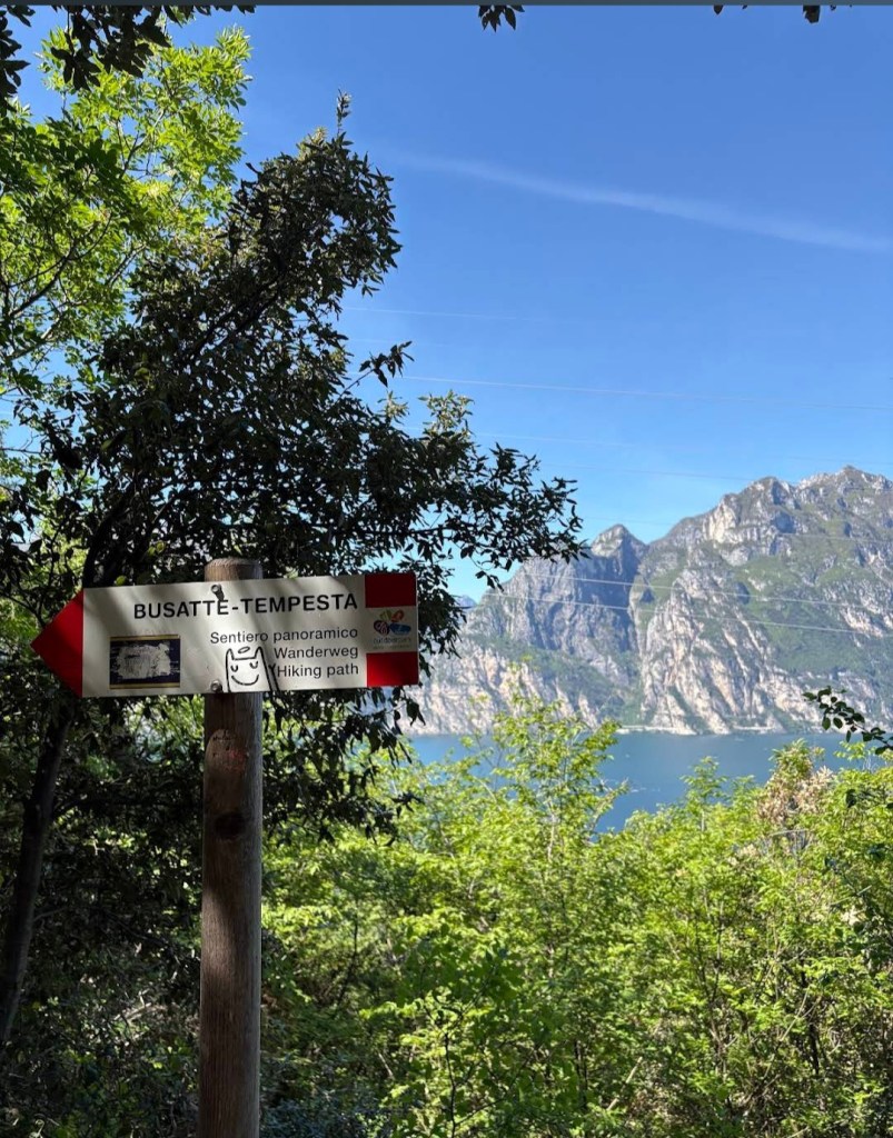Wegweiser zum Busatte-Tempesta Wanderweg oberhalb von Torbole mit Blick durch Bäume auf den Gardasee und die Berge im Hintergrund.